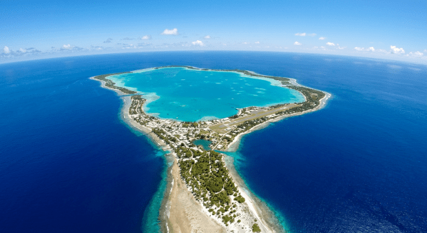 A woman in a beige trench coat stands on a grassy cliff by the ocean with rocky cliffs in the background